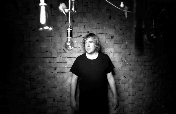 Schwarzweiß-Foto des Musikers Domikin Eulberg. Er steht in einem dunklen Raum vor einer quadratischen Holz- oder Kachelwand. Um ihn herum hängen mehrere Glühbirnen von der Decke, einige unscharf im Vordergrund. Er trägt ein schwarzes T-Shirt und blickt leicht zur Seite.