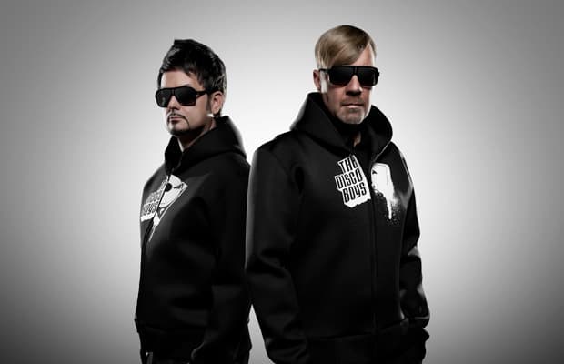 Raphael Krickow und Gordon Hollenga stehen Rücken an Rücken und blicken seitlich in die Kamera. Beide tragen schwarze Kapuzenpullover mit dem Schriftzug ‚The Disco Boys‘ und schwarze Sonnenbrillen.