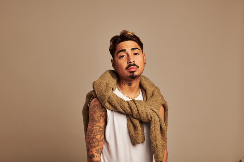 Porträt des Comedy-Artists Tony Bauer vor beigem Hintergrund, mit ärmellosem weißen Shirt, Pullover über den Schultern und sichtbaren Tattoos.